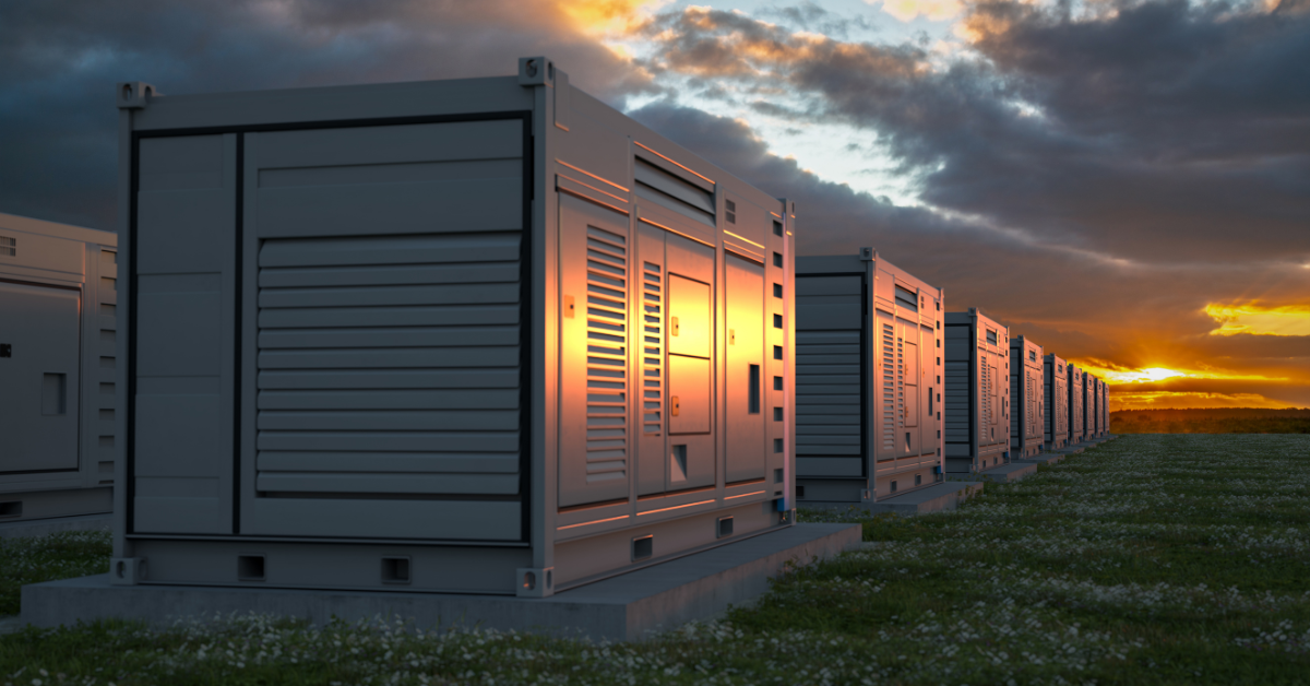 Utility-scale battery energy storage system (BESS) enclosures lined up in a grassy field at sunset, highlighting renewable energy site selection and grid interconnection capacity land requirements.